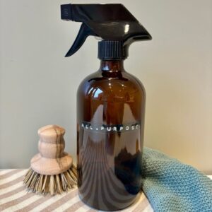 Glass amber spray bottle labelled 'All-Purpose', sat on stripey tea towel, green cloth and wooden scrub brush.