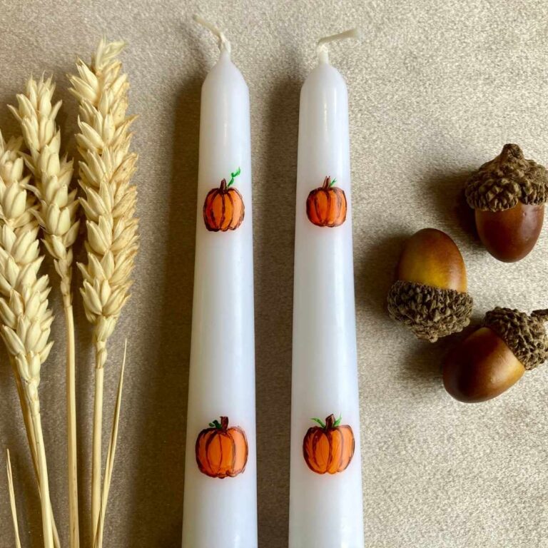 Pumpkins painted on candles next to wheat and acorns.