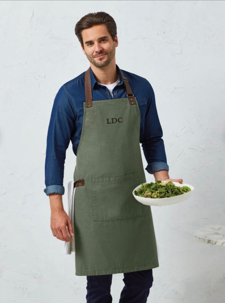 Man wearing green apron with initials on the front, holding a bowl of salad.