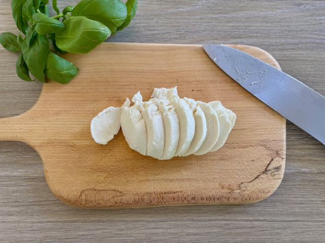 Mozzarella slices on a chopping board next to a knife and a bunch of fresh basil leaves
