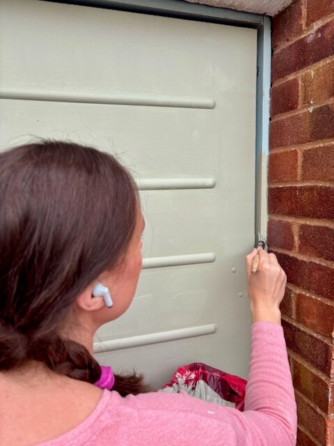 Woman using a paint brush to apply paint around the frame of the garage door.