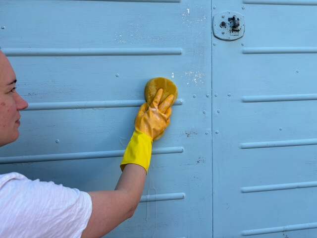 Gloved hand using a sponge to apply the sugar soap to the garage door