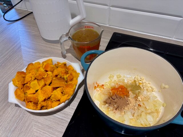 Roasted pumpkin in a white bowl, vegetable stock in a jug and Dutch oven with onions, garlic and spices in.